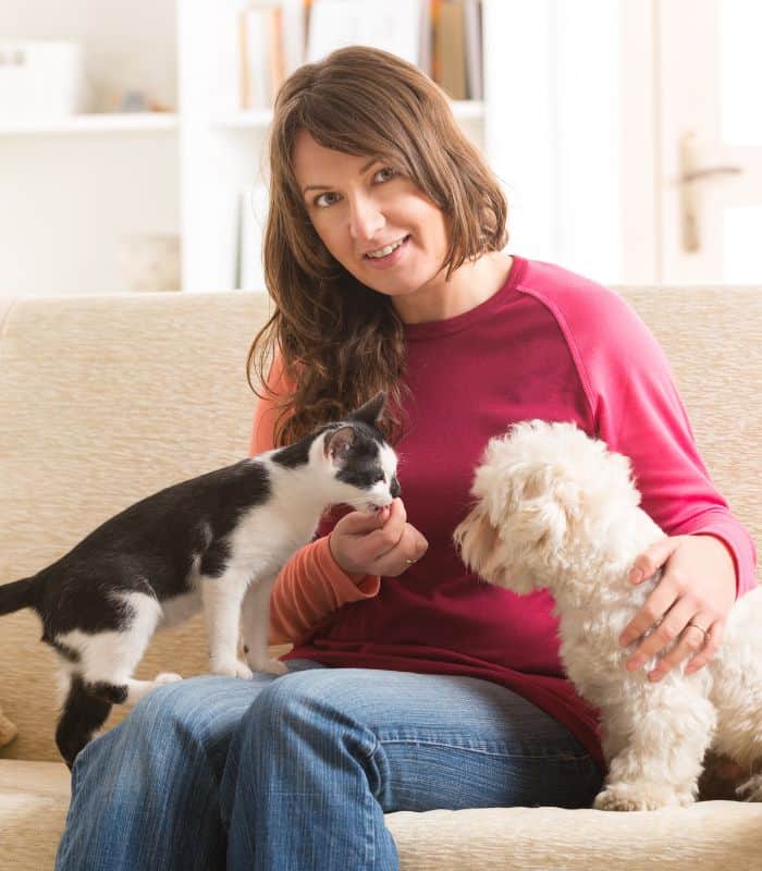 Smiling woman on couch feeding a kitten and small dog